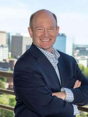 Professional portrait of a person smiling at the camera, standing outdoors with a city skyline in the background, wearing a dark blue blazer and checkered shirt.