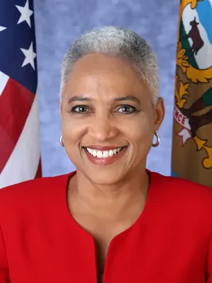 Formal portrait of a person smiling at the camera, wearing a bright red top and gold hoop earrings, with the U.S. flag and a state seal flag displayed in the background.