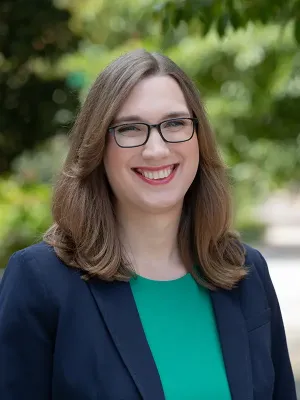 Professional outdoor portrait of a person smiling at the camera, wearing dark-framed glasses, a teal top, and dark navy blazer, with soft green foliage blurred in the background.