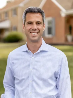 Professional portrait of a person smiling at the camera, wearing a light blue checkered shirt, standing outdoors in front of a brick building with white trim and columns.